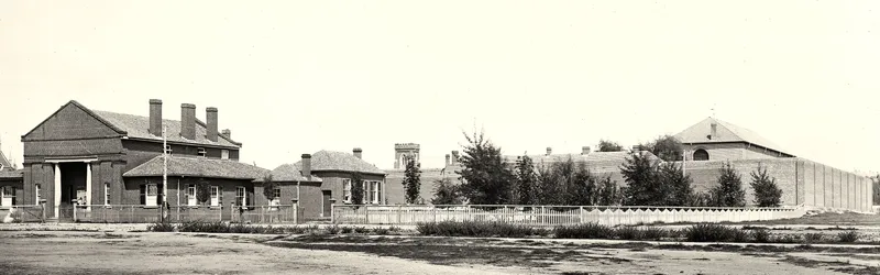 Wide photograph of a brick court house with tall chimneys and lower buildings, with the high walls of the gaol behind, beside a broad dirt road planted with young trees.