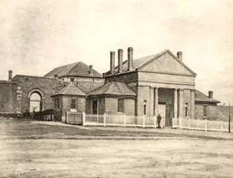 Black and white photograph of a colonial court house with a columned portico, tall chimneys and a white picket fence, set back from a wide unmade street.